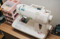 Dikiş makinesi üzerinde iplik yönü, iğne kaldırma, geri dikiş ve dikiş ayar sembolleri ile kullanım rehberi.