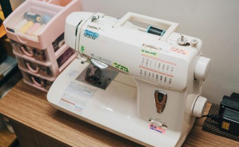 Dikiş makinesi üzerinde iplik yönü, iğne kaldırma, geri dikiş ve dikiş ayar sembolleri ile kullanım rehberi.